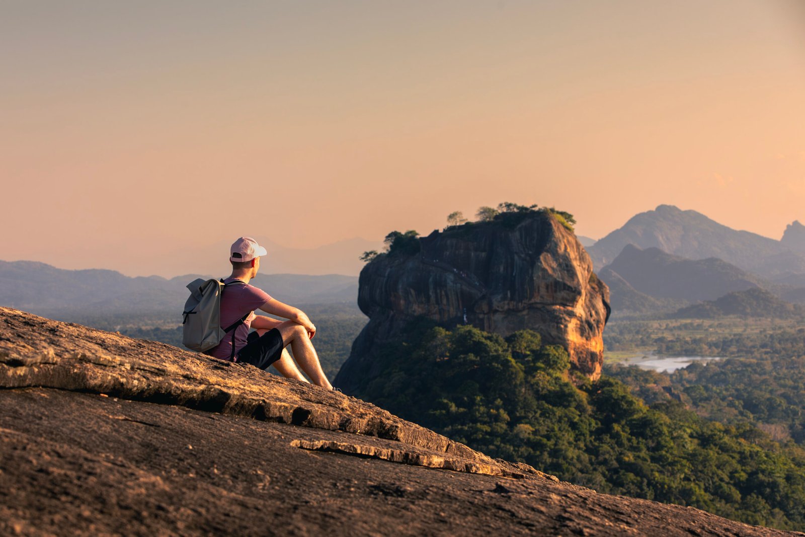 Man with backpack sitting on rock and looking at landscape at sunset. Beautiful scenery with Sigiriya rock. Solo traveler in Sri Lanka.