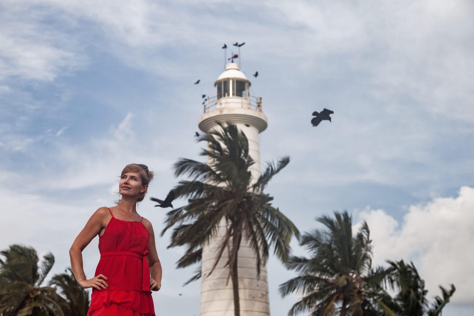A woman in a vibrant red dress stands confidently near a historic lighthouse in Sri Lanka, surrounded by tall palm trees and a dramatic sky, perfectly capturing the essence of adventure and beauty.